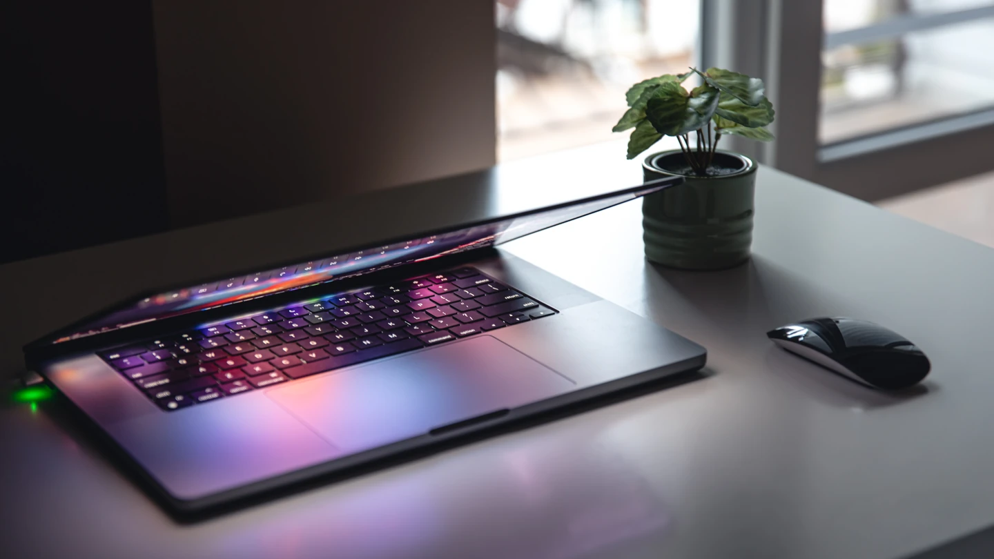 Halfgesloten laptop met verlicht toetsenbord op een bureau naast een plant en een muis